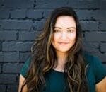 A captivating portrait of a woman smiling confidently in front of a brick wall, showcasing her long brunette hair and vibrant eyes.